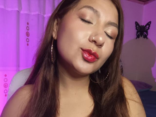 AradiaX with closed eyes, red glossy lips, nose ring, and hoop earrings, has light brown skin and long brown hair. She's pouting slightly. Purple lighting in background.