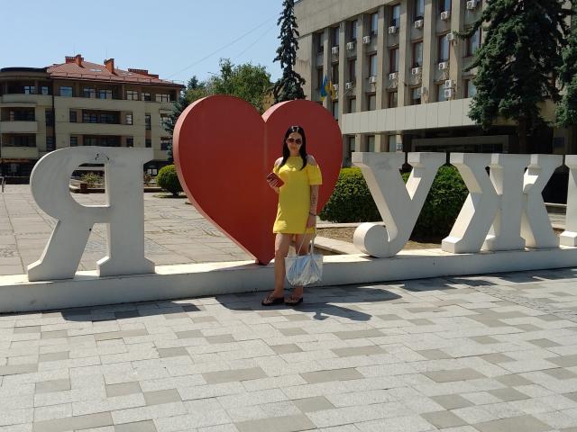 MirandaHott69 stands in front of a giant red heart and white "LOVE" letters, wearing a yellow dress, sunglasses, and sandals, holding a white purse, with a clear sky.