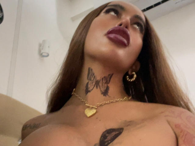 SanyaCarolina with long brown hair, dark lipstick, gold hoop earrings, and a gold heart necklace, looking up with tattoos on her neck and chest, showing her tits.