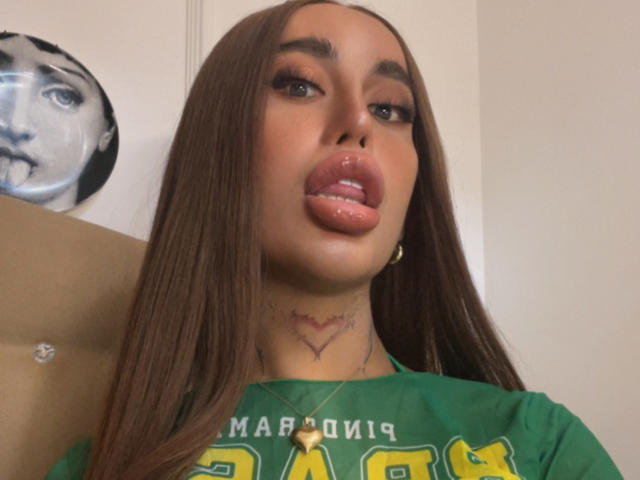 SanyaCarolina has long brown hair, full pink lips, and a tattoo below her chin. She's wearing a green shirt, and has a gold necklace. Her tits are visible.