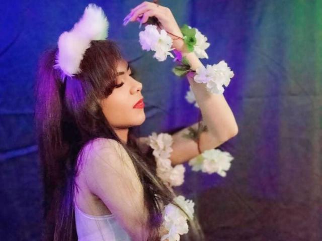 BritanyWilly with long black hair, white fluffy cat ears, and flower wristbands, eyes closed, red lipstick, raising arms, wearing a white top, blue and purple lighting.