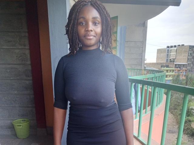 TinySnack, a dark-skinned woman with braided hair, wearing a black, form-fitting dress, standing on a balcony with green railings, looking directly at the camera, smiling slightly.
