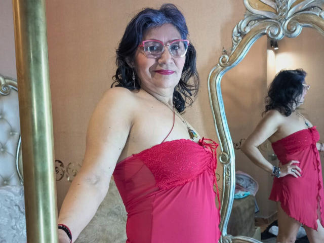 CelesteBakers with purple hair, glasses, and red lingerie, showing her tits in a mirror, hands on hips, smiling, reflection visible, gold mirror, pink wall, red lipstick.