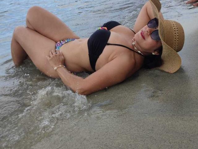 TamaraFoxy is lying in shallow water, wearing a black bikini, straw hat, and sunglasses. She's got tits, colorful panties, and a pearl necklace. Her ass is partly submerged.