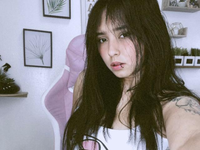 SarahMoon69 with long black hair, white tank top, pink chair, tattoo on right shoulder, slightly parted pink lips, looking at camera, white wall with framed plant art.
