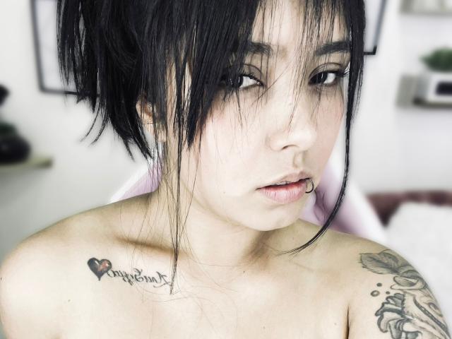 SarahMoon69 with black hair, nose ring, and tattoos on her shoulder and upper chest, looking at the camera with a slight pout. She's topless, showing her tits.