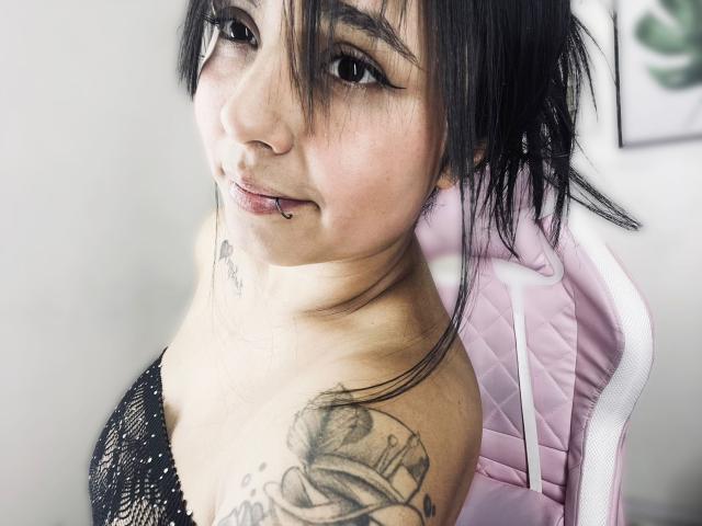 SarahMoon69 with black hair, eyeliner, nose ring, and lip piercing, showing tattooed shoulder, wearing black lace top, sitting in pink chair, smiling slightly.