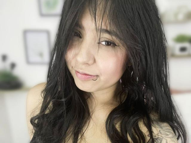SarahMoon69 has long, black, wavy hair, a cute nose ring, and is looking at the camera with a slight smile. She has light skin and brown eyes.