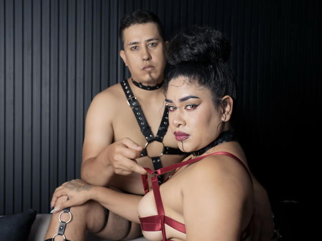 LianaAndAsaff with dark hair in a bun, wearing red harness and black choker, has her tits pointed at by Asaff with black hair, wearing black harness and choker.