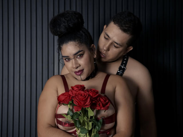LianaAndAsaff with dark hair in a bun, wearing red rose-patterned lingerie, holding red roses, has a muscular man with a topknot, black choker, leaning behind her.