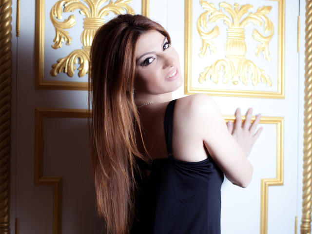 Sexydollhotx avec de longs cheveux bruns, un haut noir sans manches, regardant en arrière, souriant, tenant une porte avec des motifs dorés ornés, un mur blanc et doré, montrant les épaules nues et le haut du dos..