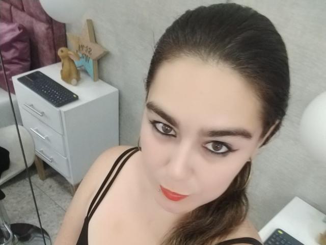 TaylorNova, com cabelo castanho escuro preso em um rabo de cavalo, vestindo lingerie preta, tem batom vermelho, delineador preto e está olhando para a câmera com um leve sorriso. Seios visíveis..