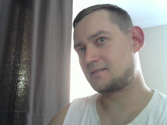 CharlieYeah with a shaved head, stubble, and a white tank top, looking slightly confused, standing near a dark textured curtain.