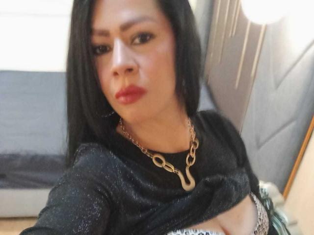 SofiMontes, com cabelo escuro, batom vermelho e uma blusa preta brilhante, mostrando um pouco dos seios, usando um colar de corrente dourada, em um selfie borrado..