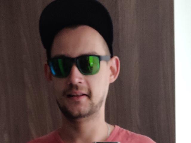 CharlieYeah has a black cap, green-tinted sunglasses, and a pink shirt. He's slightly smiling with a short beard and a brown wood background.