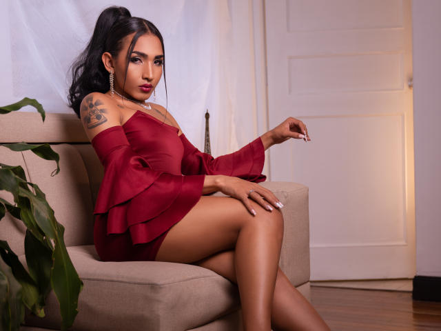 GabySantos with dark hair in a ponytail, wearing a red off-shoulder dress, sitting on a beige chair, showing a tattoo on her right arm, and smoking a cigarette.