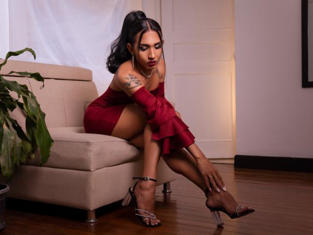 GabySantos in a red dress, sitting on a beige sofa, adjusting her silver high heels, showing her tattooed arm, with her dark hair in a ponytail, looking down.