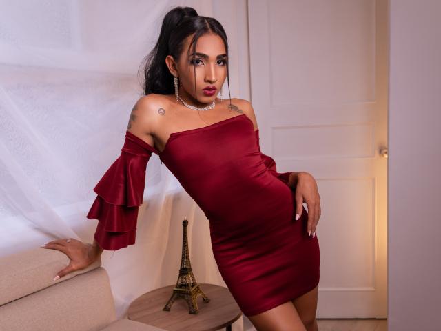 GabySantos in a red off-shoulder dress, standing with one hand on a beige couch, wearing a necklace, with tattoos on her shoulders, and a small Eiffel Tower figurine.