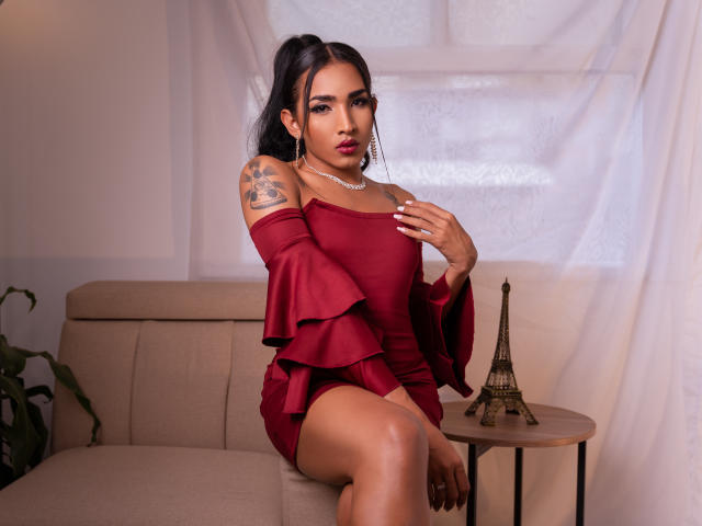 GabySantos with dark hair in a ponytail, wearing a red off-shoulder dress, showing a tattoo on her right arm, sitting on a beige couch, next to a mini Eiffel Tower.