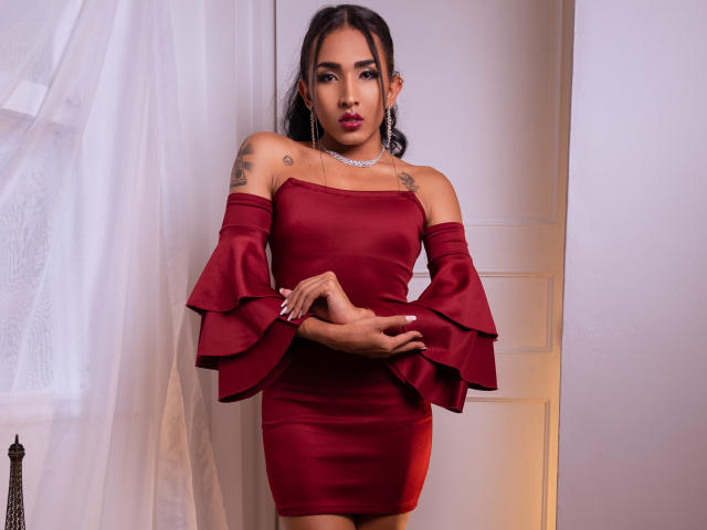 GabySantos in a red off-shoulder dress with flared sleeves, standing with arms crossed, showing tattoos on her shoulders, and wearing a silver necklace, with a confident look.