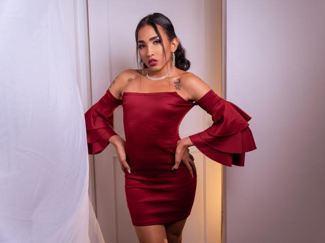 GabySantos with dark hair in a ponytail, wearing a red off-shoulder dress with flared sleeves, hands on hips, tattoos on shoulders, necklace, standing in a doorway.