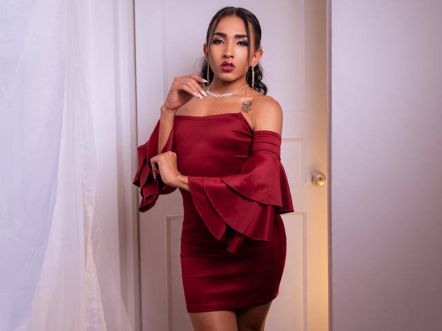 GabySantos with dark hair, wearing a red off-shoulder dress, flared sleeves, and a necklace, stands in a doorway, touching her chin, showing a small tattoo on her chest.