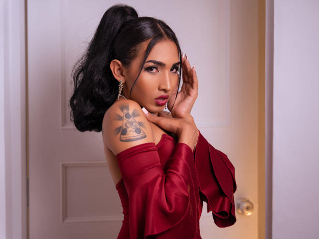 GabySantos with dark hair in a ponytail, wearing a red off-shoulder dress, hands near face, showing a tattoo on her left shoulder, with earrings and a serious expression.