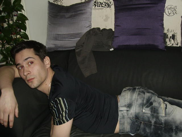Ethan69, with short dark hair and red eyes, lies on a black couch, wearing a black striped shirt and faded jeans, looking at the camera with a slightly bored expression.