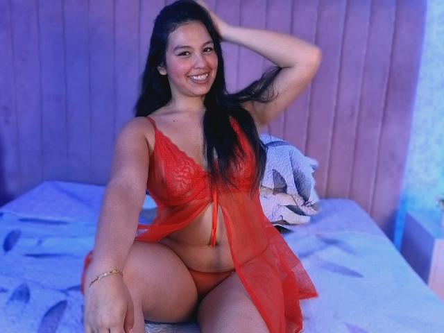 SarahXCarter with long black hair, smiling, wearing a red lace lingerie set, sitting on a bed, hand in hair, showing her red panties and lace bra, blue-tinted room.