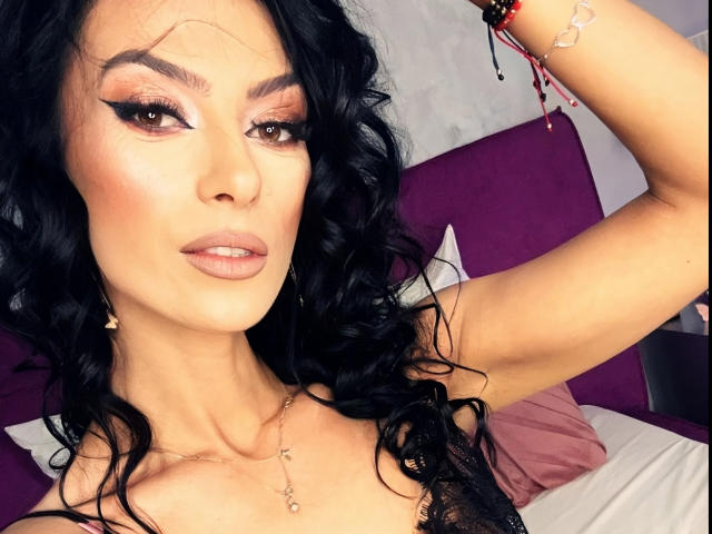 KinkyAmelie with wavy black hair, bold eyeliner, and pink lipstick, showing off her tits in a black lace top, arm raised, pink pillow in background.