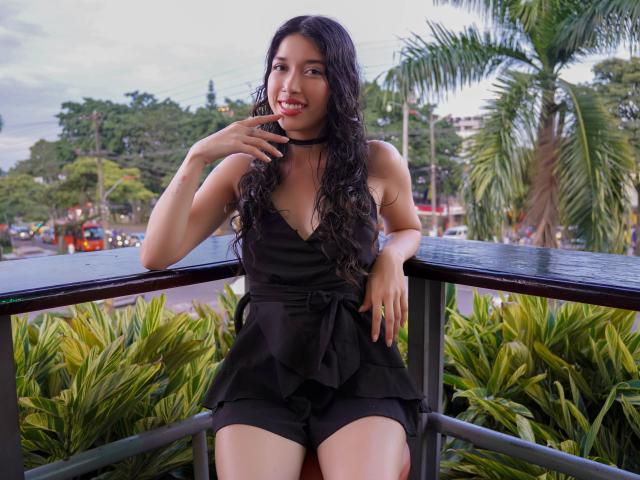 Krystal Evans, con lunghi capelli neri e ricci, indossa un abito nero con un dettaglio allacciato sul davanti, è seduta su un balcone, con un dito vicino alla bocca, sorridendo. Sullo sfondo si vedono piante verdi e una palma..