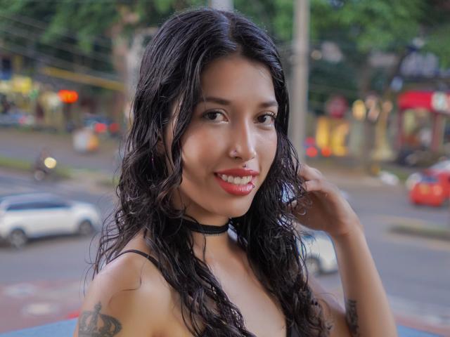 Krystal Evans, con cabello negro ondulado, lápiz labial rojo, collar negro, sonriendo, mostrando tatuajes en su brazo y tocándose el cabello. Se ven sus senos, el fondo de la calle está borroso..