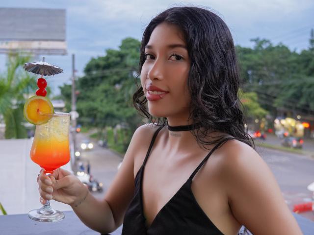 KrystalEvans with curly black hair, wearing a black halter top, holding a colorful cocktail with a lemon slice and umbrella, looking at the camera, outside at dusk.