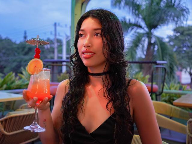Krystal Evans, com longos cabelos negros ondulados, usando um colar preto e uma blusa sem mangas, segurando um coquetel vermelho com um guarda-chuva e uma fatia de laranja, sorrindo para a câmera..