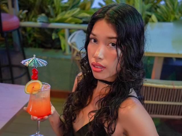 KrystalEvans with curly black hair, holding a colorful cocktail with a mini umbrella and fruit, wearing a black choker and strapless top, looking at the camera.