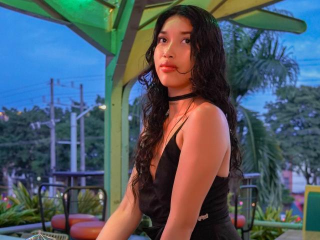 KrystalEvans with long, wavy black hair, wearing a black choker and sleeveless black dress, sitting outside at dusk, looking off to the side, with a subtle, confident expression.