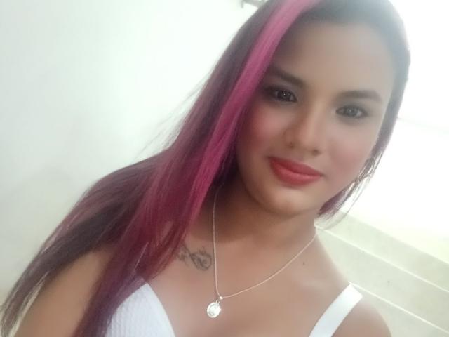 JennyLover69 con reflejos rosados, camiseta blanca, collar de plata y lápiz labial rosa, mostrando pequeños senos y un tatuaje en el pecho, sonriendo a la cámara..