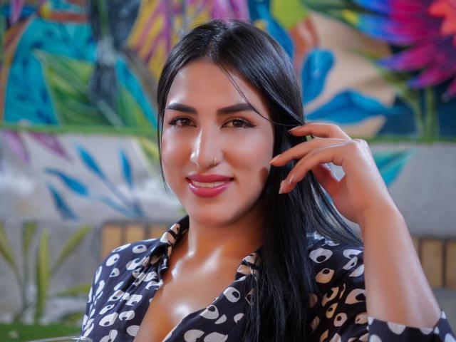 AlanaHarris with long black hair, brown eyes, and pink lipstick, wearing a black patterned shirt, smiling, hand in hair, showing her tits, in a brightly colored room.