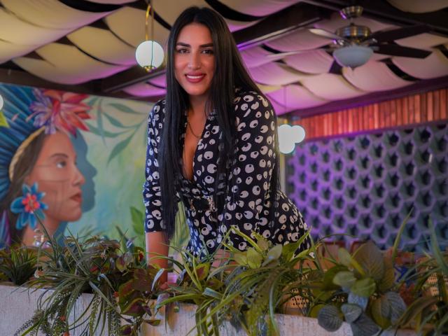 AlanaHarris, with long black hair and a smile, leans on a plant-filled ledge, wearing a black, white-patterned, low-cut dress, showing her tits.