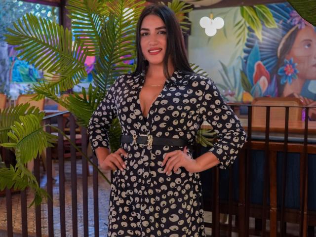 AlanaHarris with long dark hair, smiling, wearing a black dress with white eye patterns, belt, standing with hands on hips, surrounded by green plants. Her tits are visible.