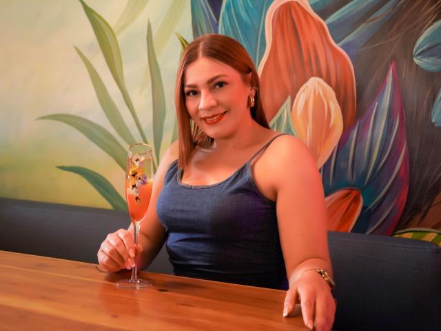 SamantaKing with red hair, blue tank top, smiling, holding a tall orange cocktail with star decorations, sitting at a wooden table, in a colorful, tropical-themed setting.