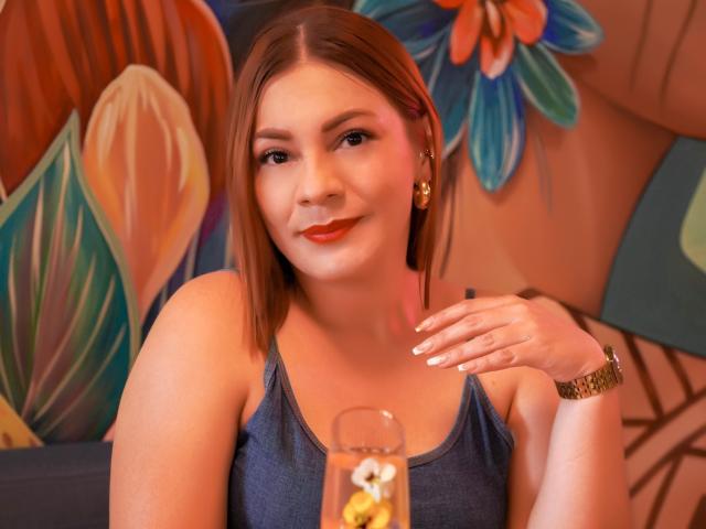 SamantaKing with red lipstick, blue tank top, and gold earrings, holding a glass with a lemon, smiling softly, right hand near face, brown hair, gold watch, warm lighting.