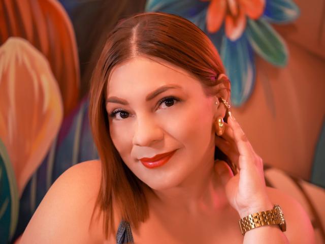 SamantaKing with red lipstick, brown hair, gold earrings, and a gold watch, smiling softly, touching her ear, wearing a blue top, looking cute.