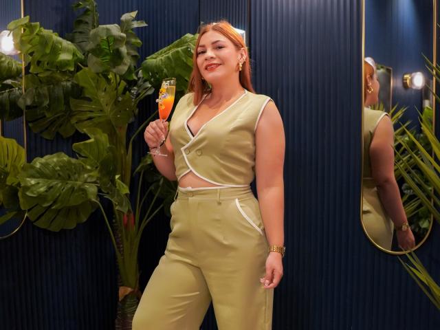 SamantaKing with red hair, green outfit, holding a champagne flute, smiling, showing her tits in a sleeveless top, standing by a plant, reflected in a mirror.