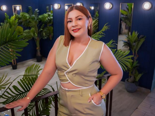 SamantaKing with red hair, smiling, wearing a beige vest with white trim, exposing her tits, hands in pockets, gold watch, standing by a railing with green plants.