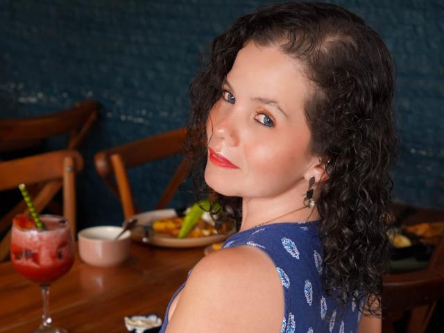 MadiFoxy with curly dark hair, blue patterned dress, red lipstick, smiling at camera, looking over shoulder, red drink with straw on wooden table, blue textured wall background.