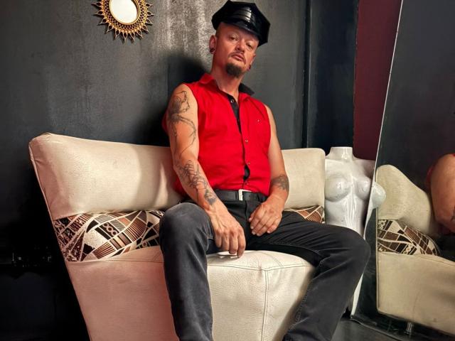 ZainKreiger, tattooed, wearing a black leather cap, red sleeveless shirt, black pants, sitting on a beige couch with geometric pillows, showing a confident, sexy, dominant look.