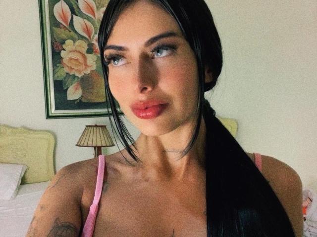 SaraCherryy with black hair in a ponytail, wearing a pink top, has shiny red lips and blue eyes, with a tattoo on her right arm, looking to the side.