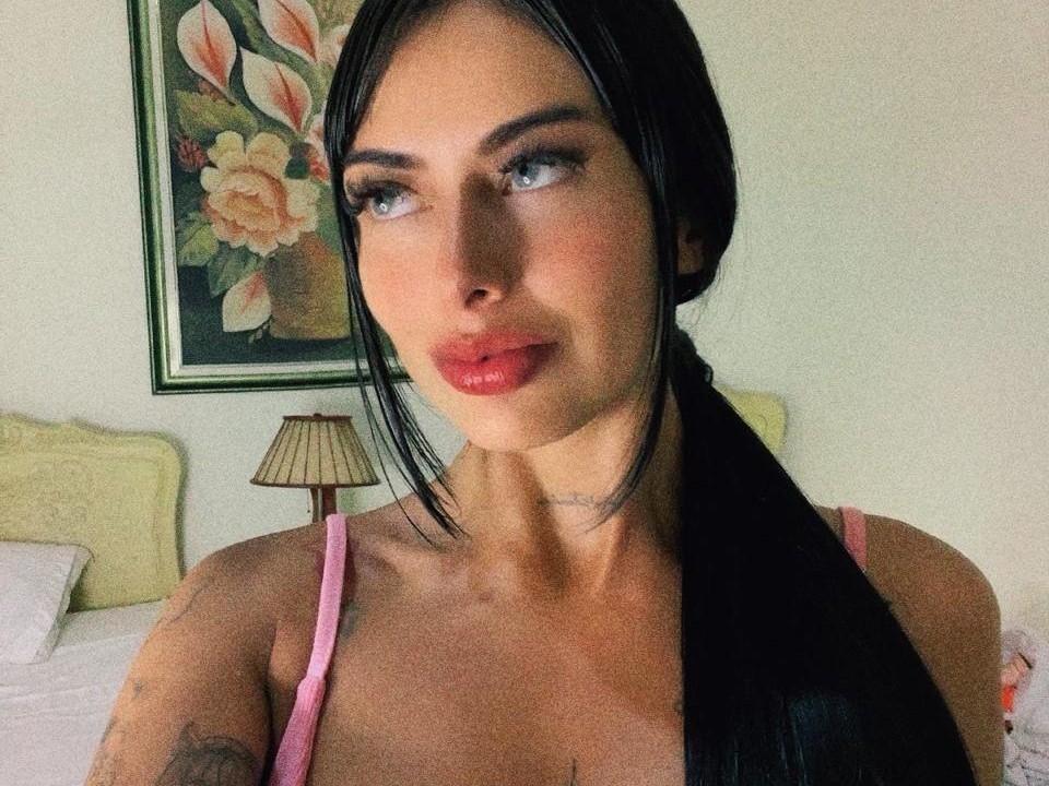 SaraCherryy with black hair in a ponytail, wearing a pink top, has shiny red lips and blue eyes, with a tattoo on her right arm, looking to the side.