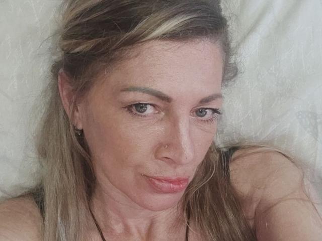 Het gezicht van CharlenePearl, met lichtbruine ogen, blond haar met een vlecht aan de zijkant, een klein neuspiercing, een lichte frons en subtiele make-up, ligt op een wit kussen..